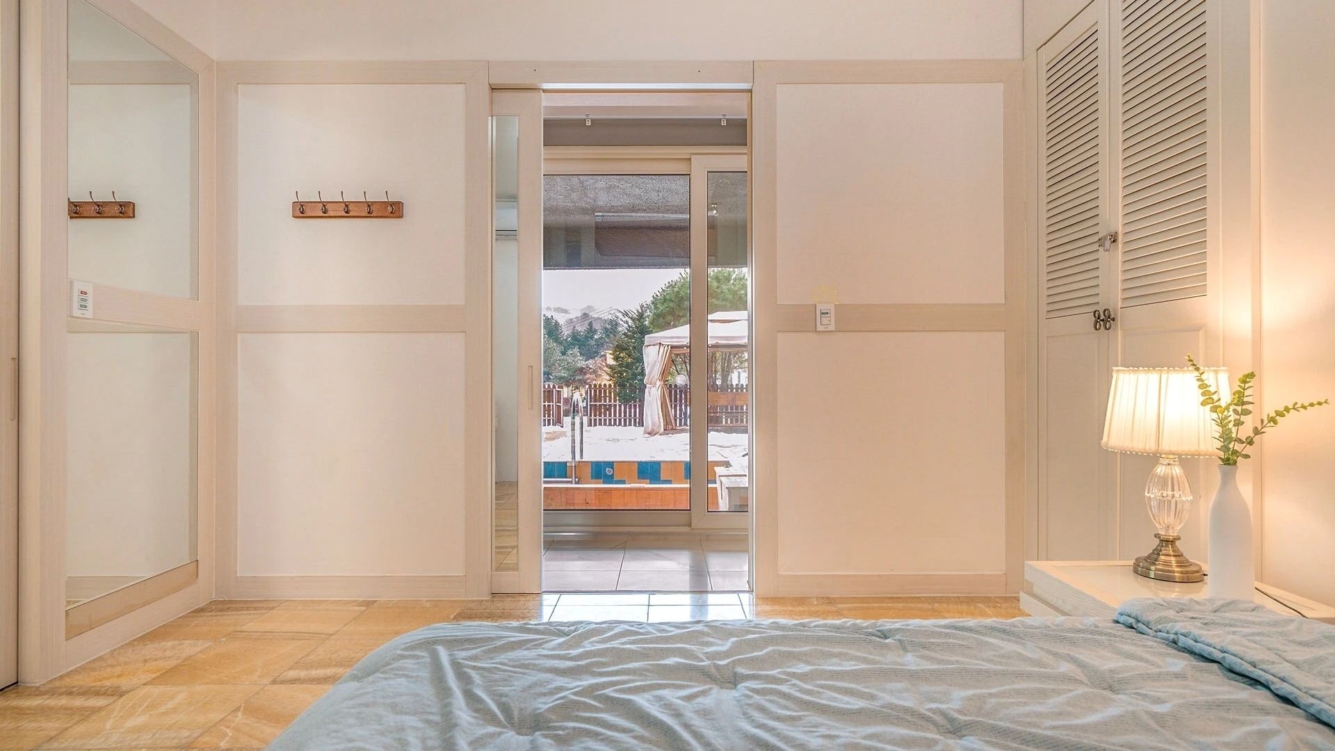 Un dormitorio con cama, una lámpara de noche y vistas a un área exterior con piscina y valla.