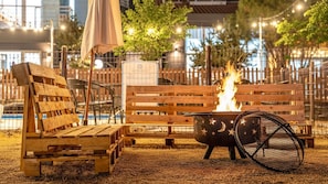 Un banc en bois, un foyer et un barbecue en métal, avec une clôture et des arbres en arrière-plan.