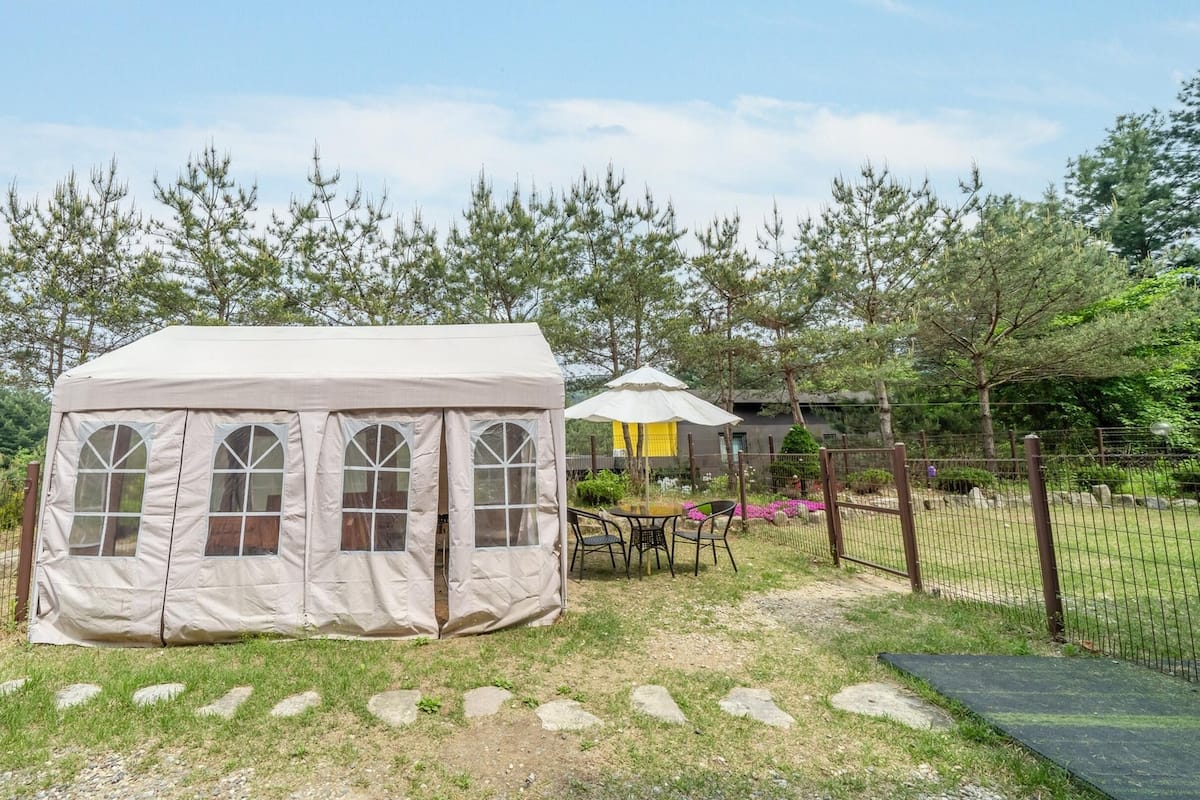 A tent with arched windows, a table with chairs, and a fenced garden area with trees and a building in the background.