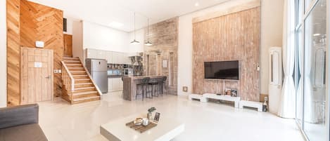 A modern living room with a flat-screen TV, a coffee table, and a staircase leading to a kitchen area.