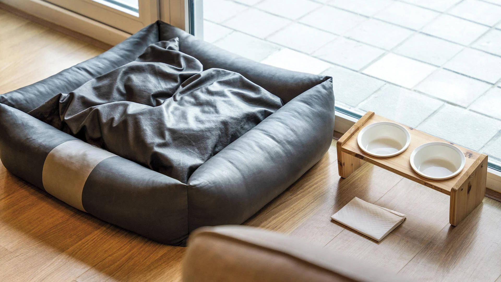 Una cama para mascotas con cojín, un comedero de madera con cuencos y una ventana con vistas al exterior.