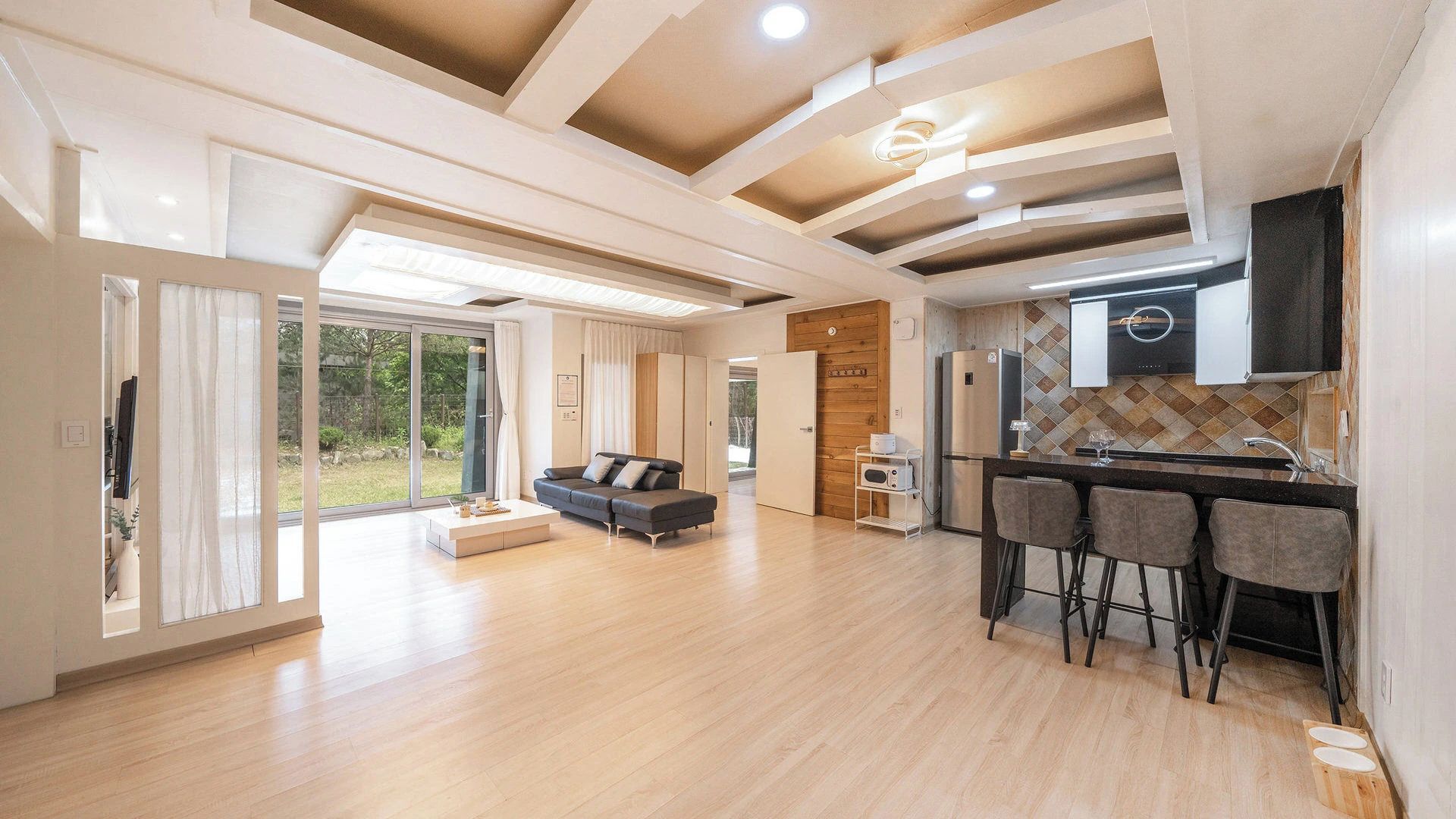 Un amplio salón con cocina moderna, una zona de comedor con taburetes de bar y un gran ventanal corredero que da a un jardín.