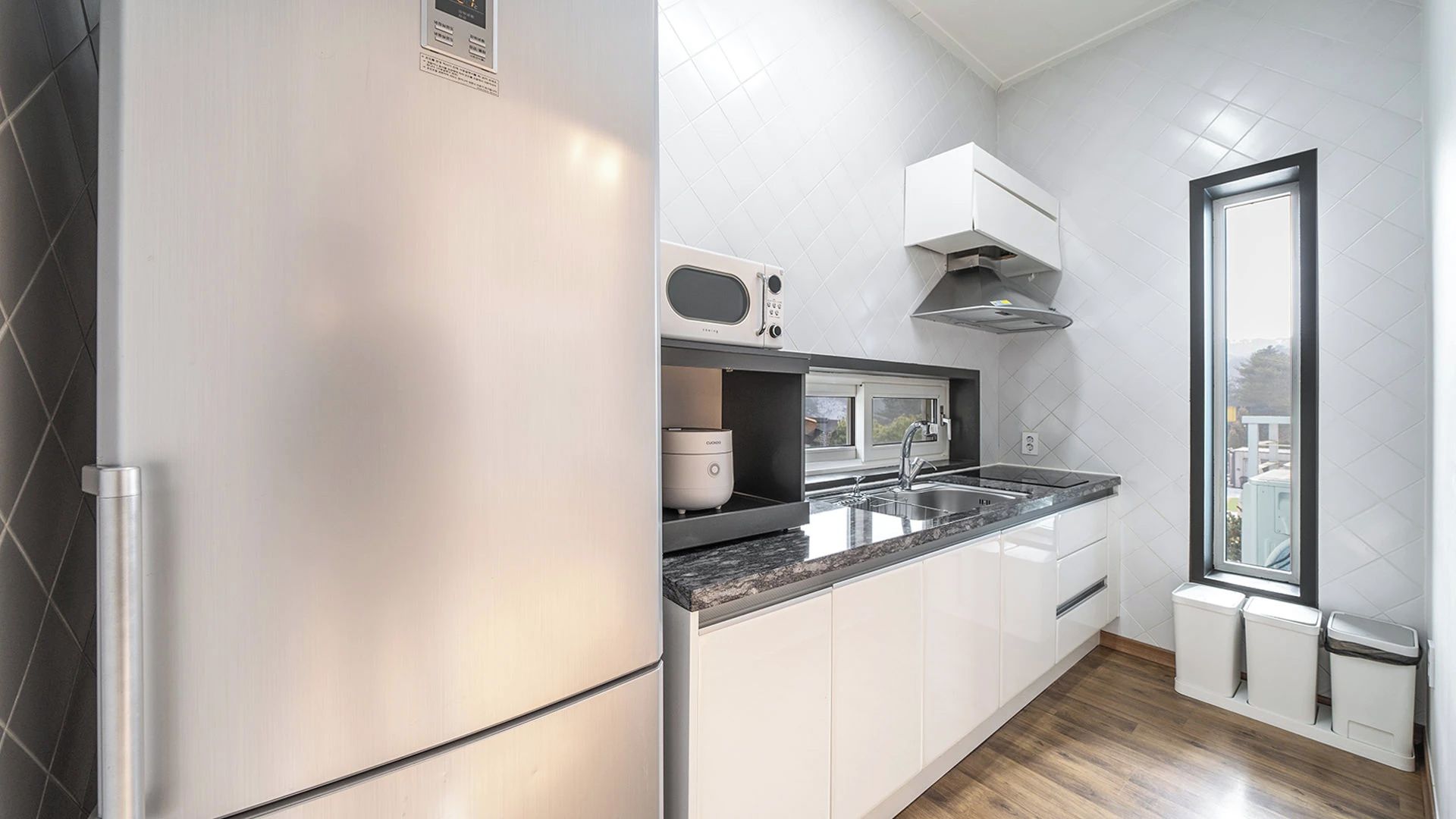 A modern kitchen with a stainless steel refrigerator, a microwave, and a built-in oven. There is a window providing a view of the outdoors.