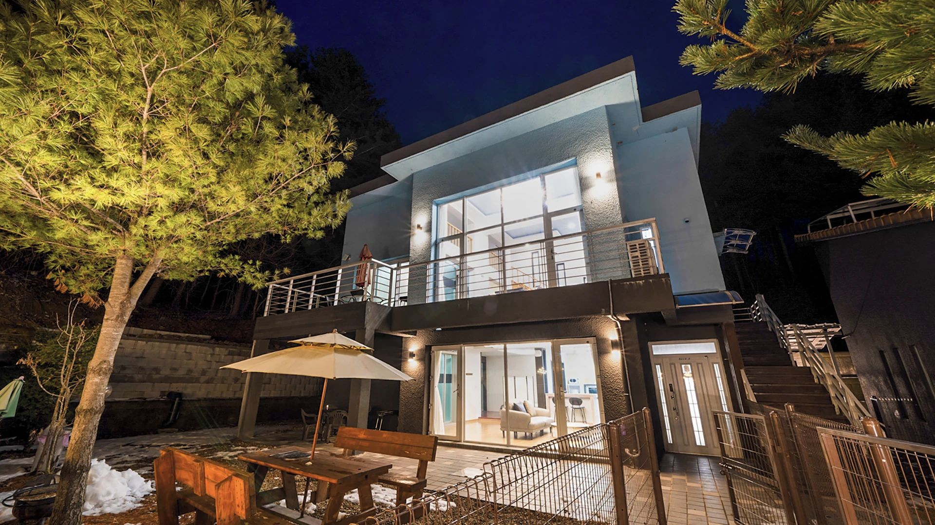 A modern two-story house with a balcony, a patio with a table and chairs, and a staircase leading to the upper level.
