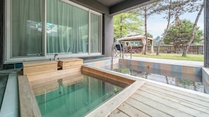 A hot tub with wooden steps, a window with curtains, and a deck area with outdoor furniture.