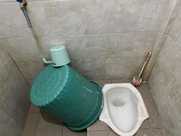 Une petite salle de bain avec des toilettes à la turque, un récipient d’eau vert et une brosse pour les toilettes.