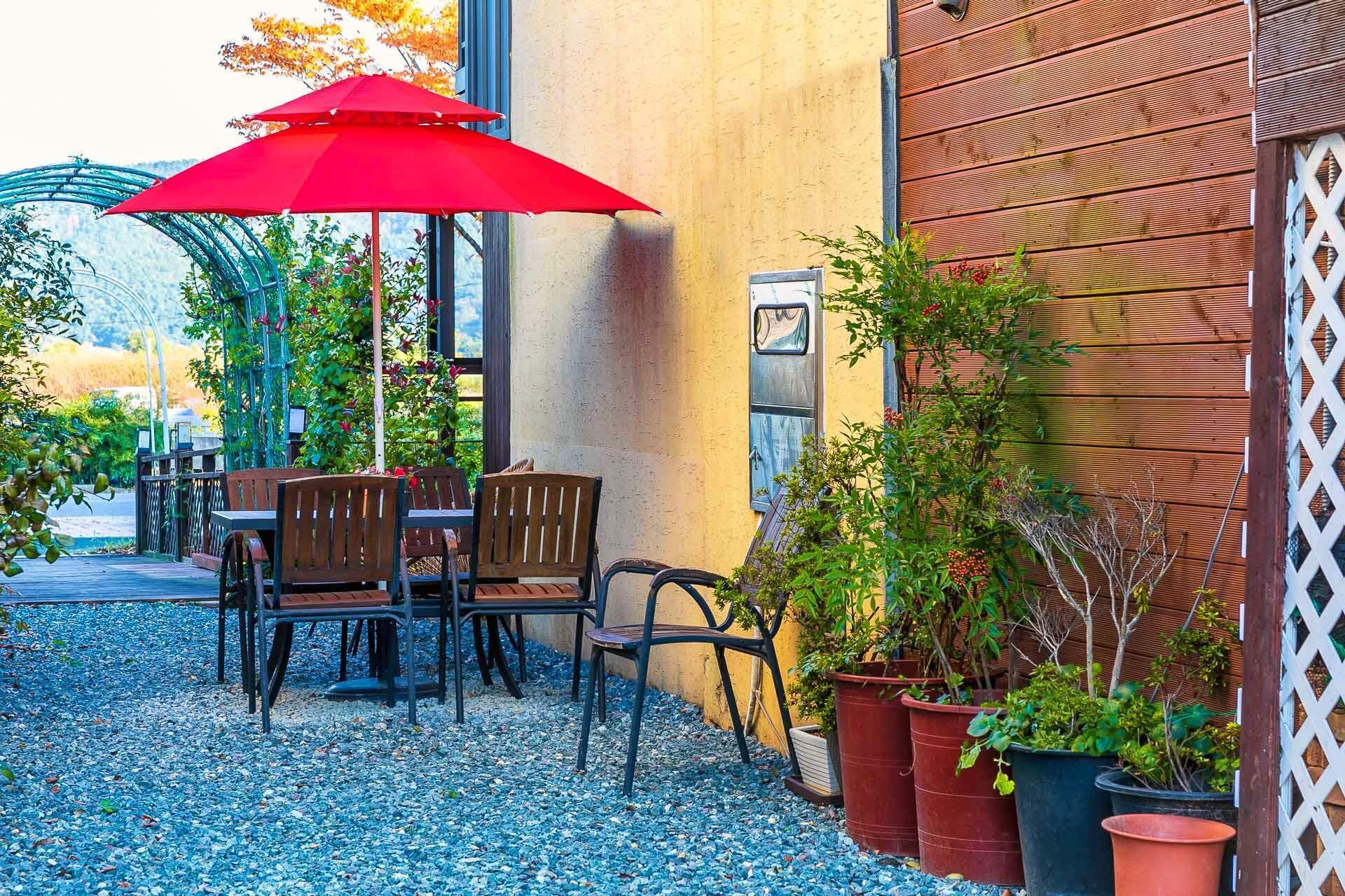 Patio esterno con tavolo e sedie, un ombrellone rosso e piante in vaso.
