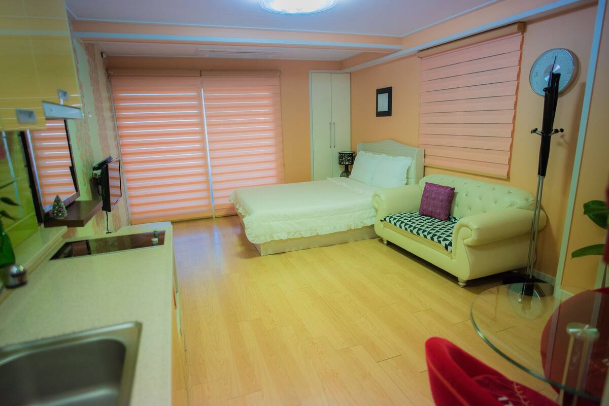 A room with a bed, a sofa, a small table, a mirror, and a window with blinds.