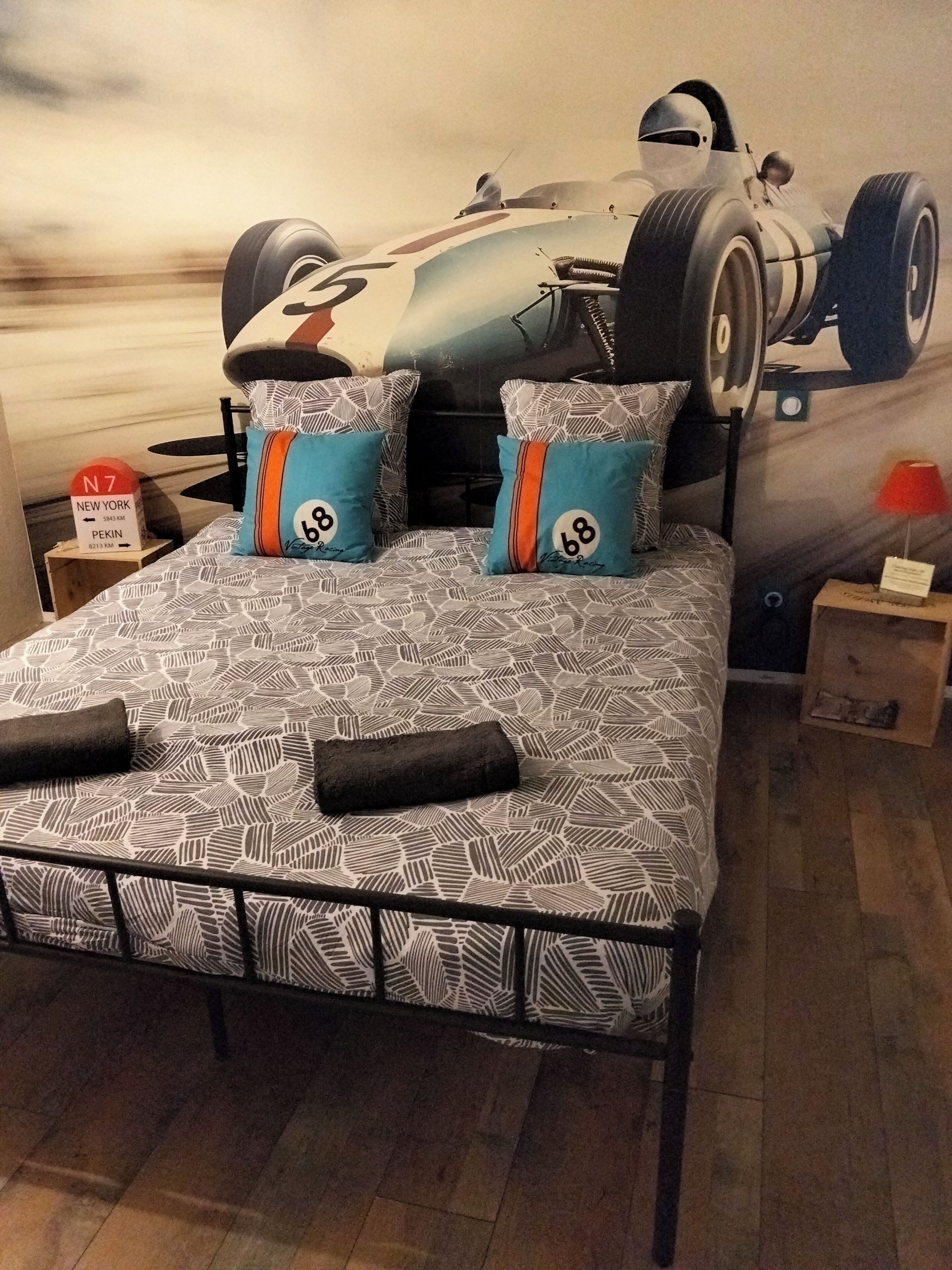 A bed with a patterned bedspread, two pillows with racing numbers, a headboard, and a wall mural of a race car.