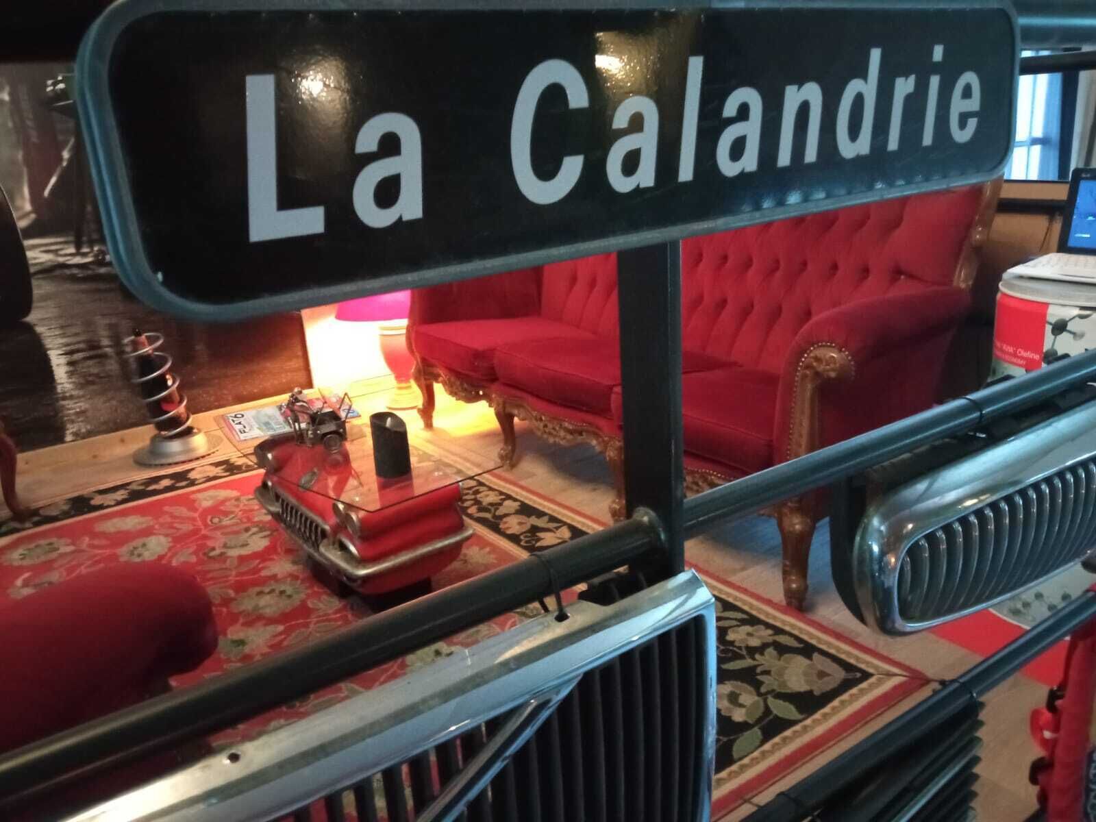 A sign reading "La Calandrie" in a room with a red sofa, a small table with a lamp, and a vintage car model.