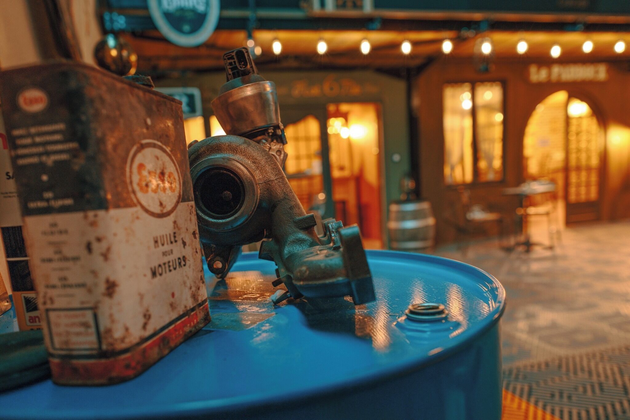 A blue barrel with a rusty can and a metal valve on top, set against a blurred background of a warmly lit interior space.