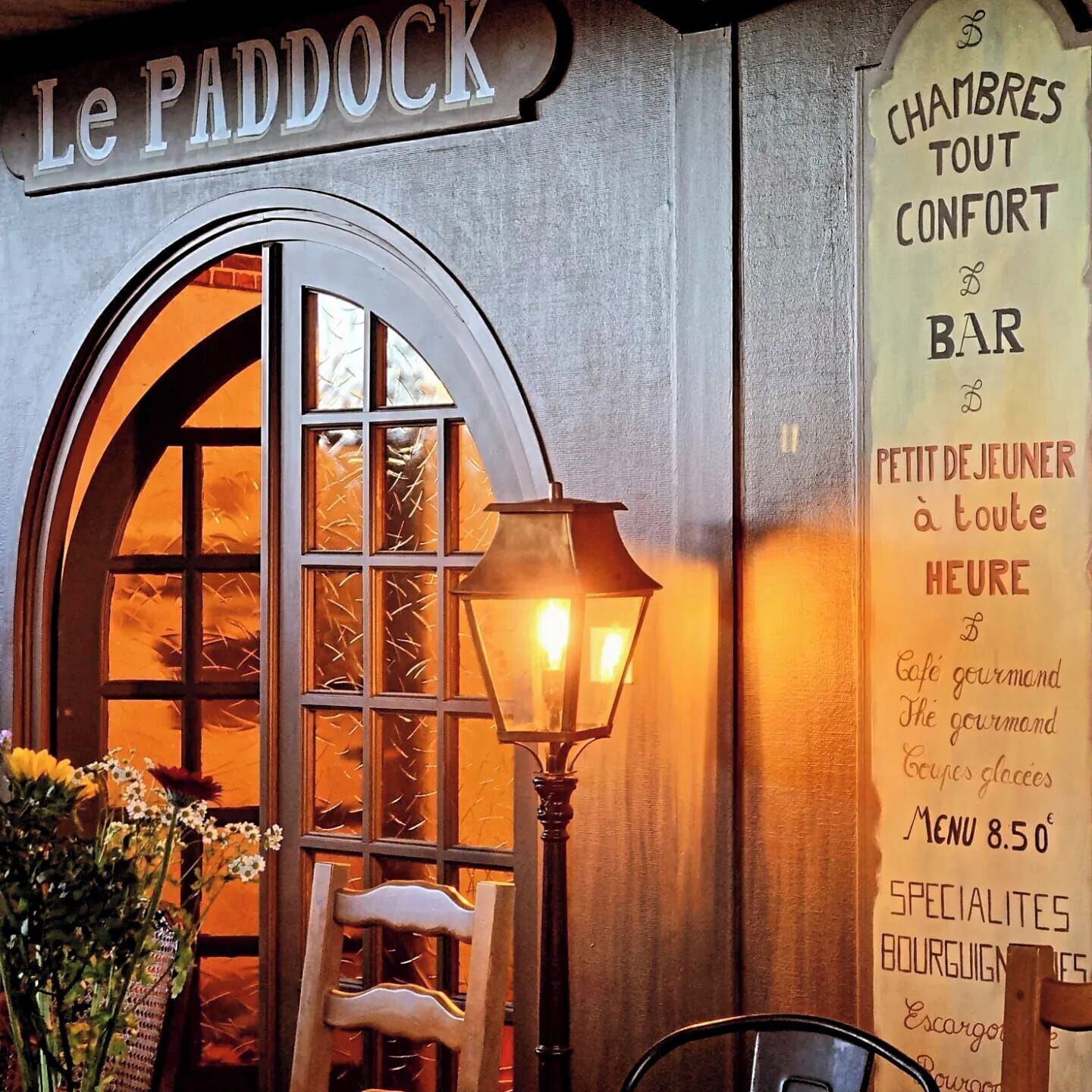A cozy interior with a sign reading 'Le Paddock' and a menu board listing 'Chambres Tout Confort Bar' and 'Petit Dejeuner à toute Heure.'