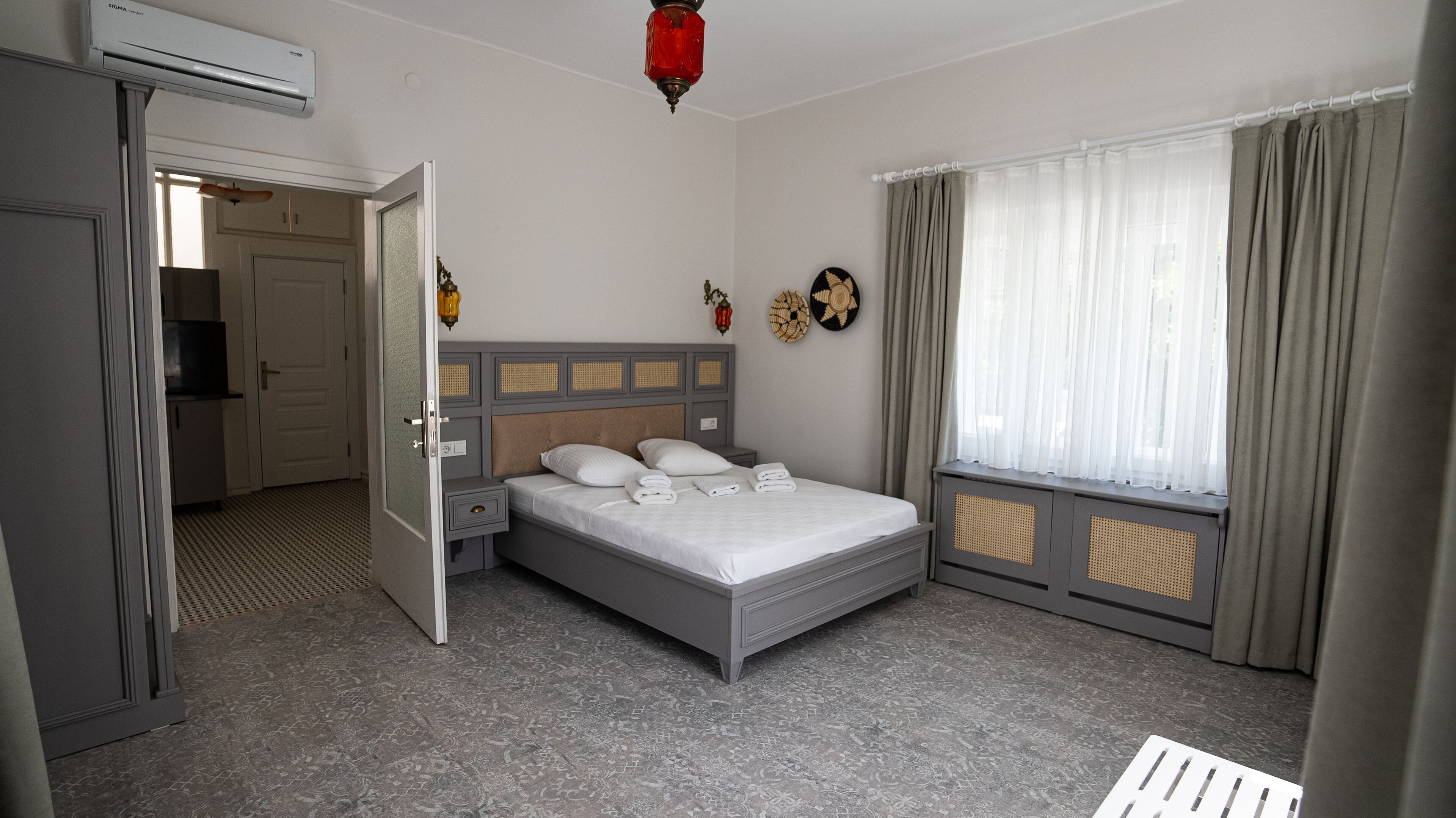 Una camera da letto con un letto, comodini, un armadio, una finestra con tende, un condizionatore d'aria a parete e una porta che conduce in un'altra stanza.