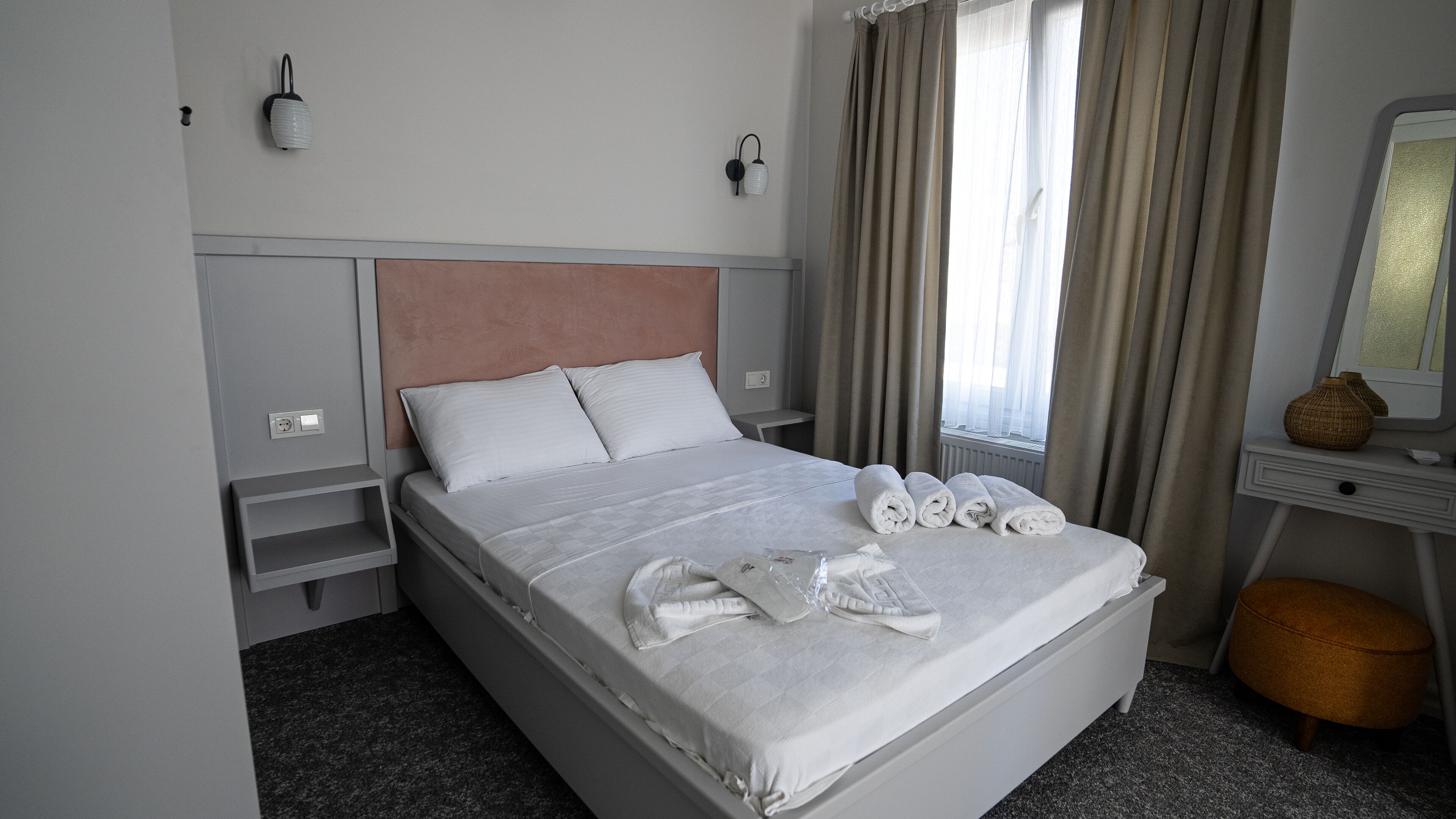 A neatly made bed with white linens and rolled towels, a bedside table with a lamp, a window with curtains, and a small bedside table with a vase.