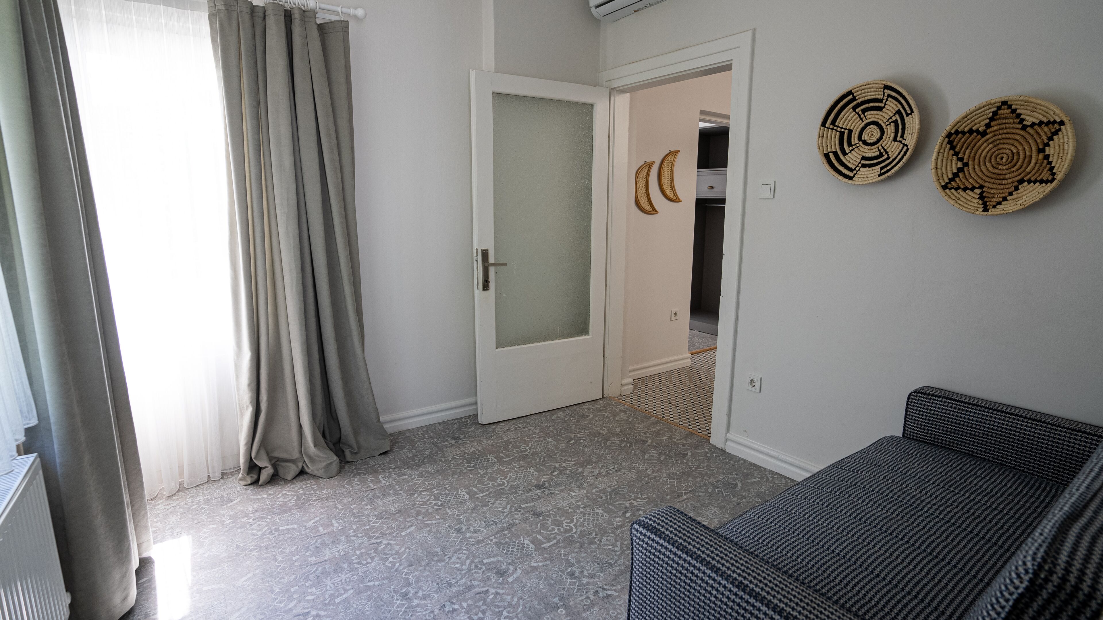 A room with a grey patterned floor, a white door with frosted glass, a grey sofa, and two woven wall decorations.