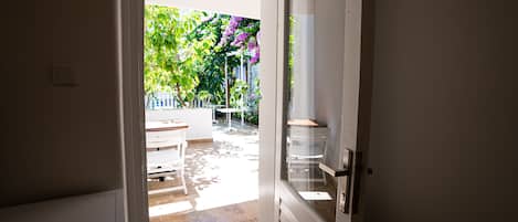 Puerta abierta que da a un patio con mesa y sillas, rodeado de vegetación y una cerca blanca.