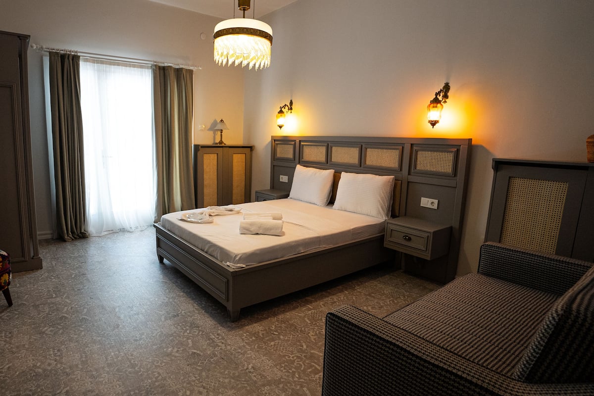 Quarto de hotel com uma cama grande, duas luminárias de cabeceira, uma cadeira, cômoda e um espelho.