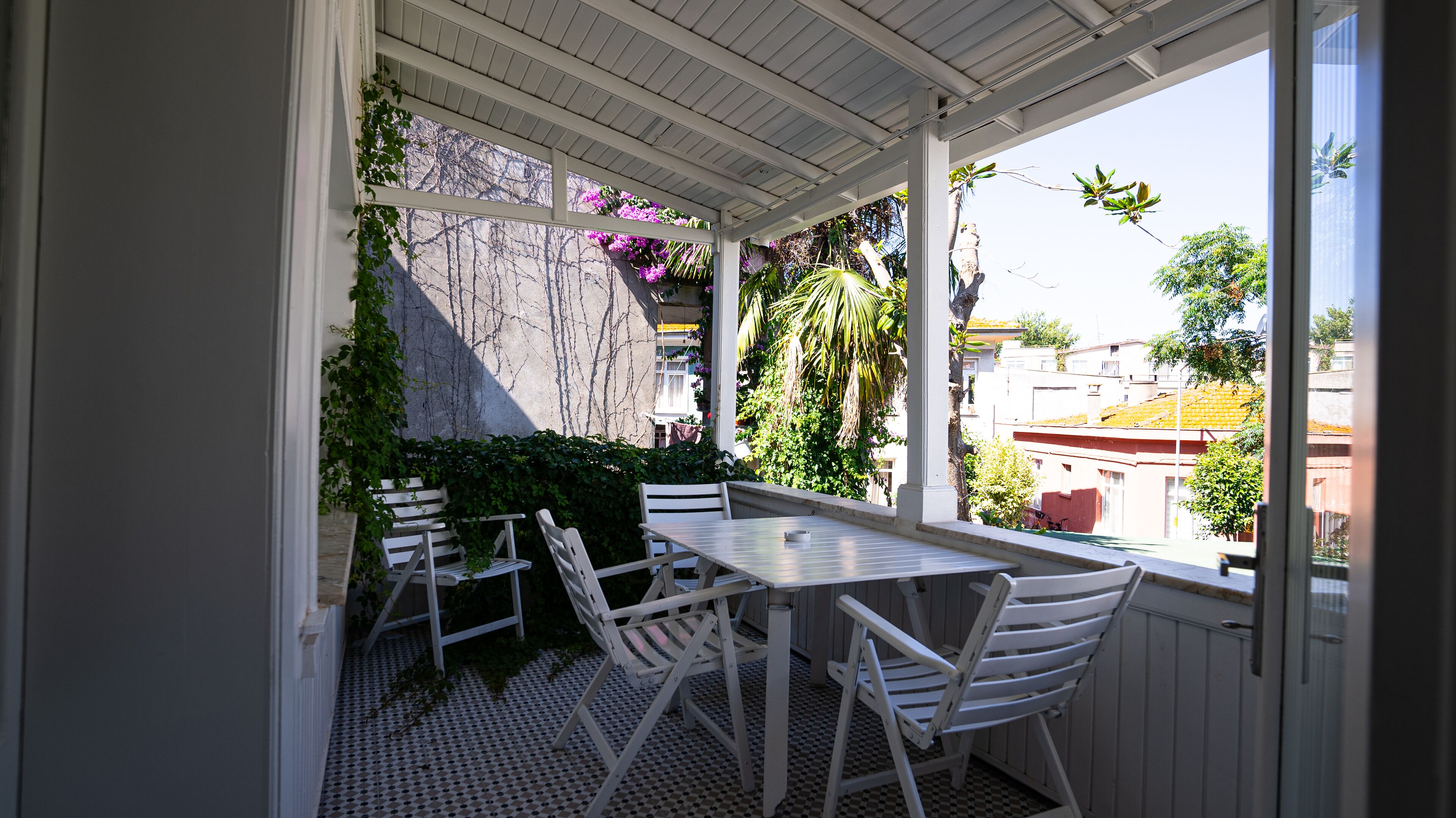 A covered patio with a table and chairs, surrounded by greenery and a view of buildings.