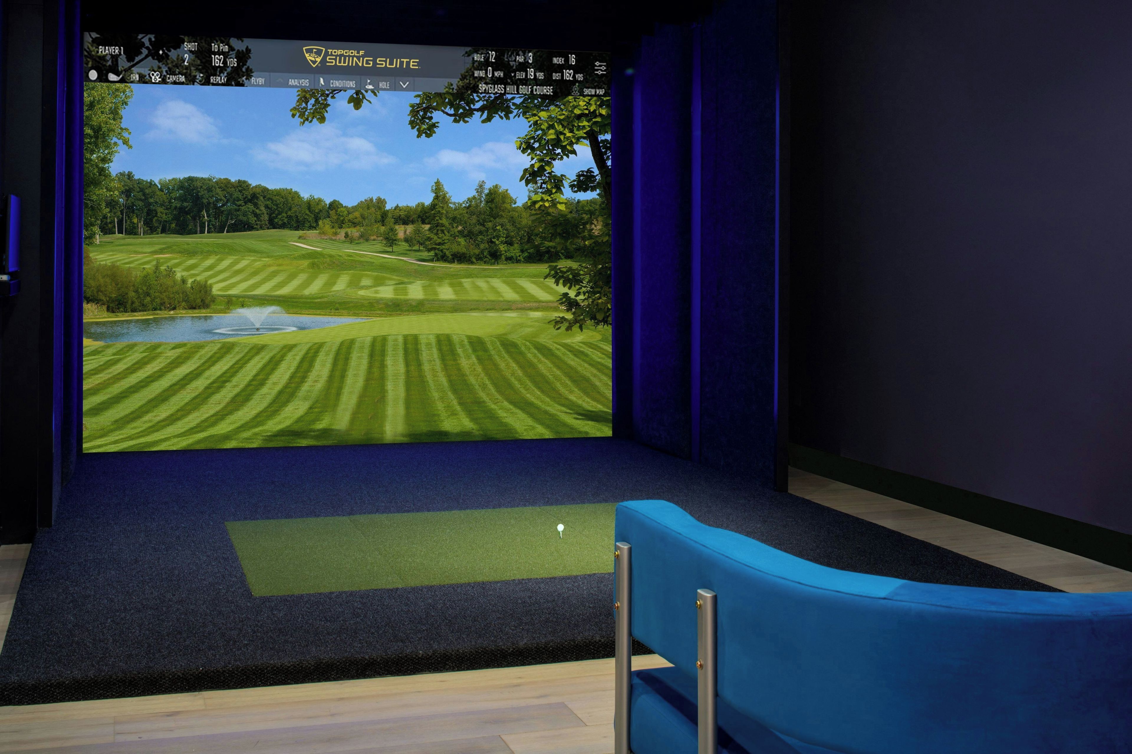 A room with a large screen displaying a golf course view, a blue chair, and a carpeted floor.