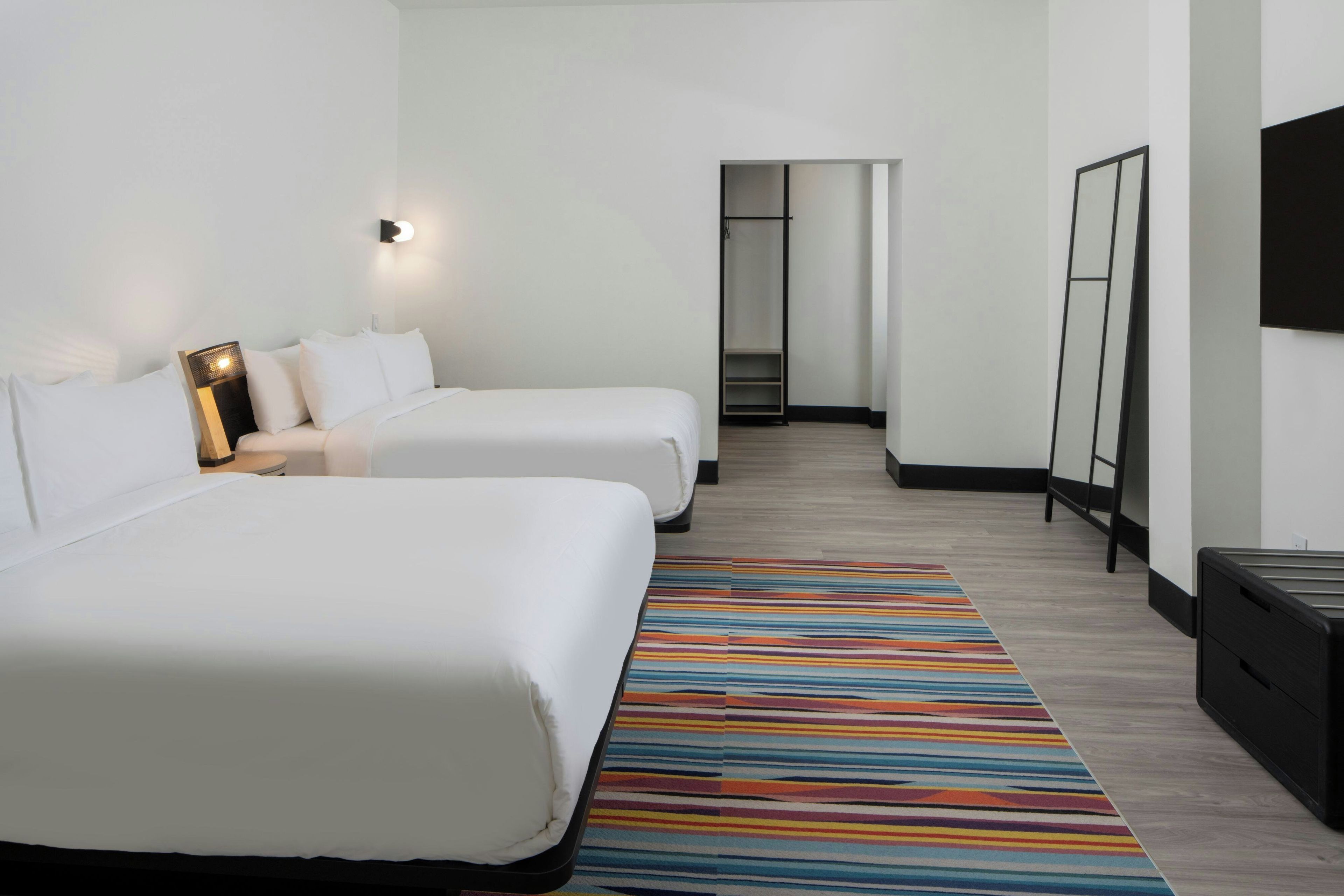 A hotel room with two beds, a striped rug, a bedside table with a lamp, and a mirror on the wall.