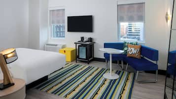A modern hotel room with a bed, a small table, a sofa, a TV, and a window with a city view.