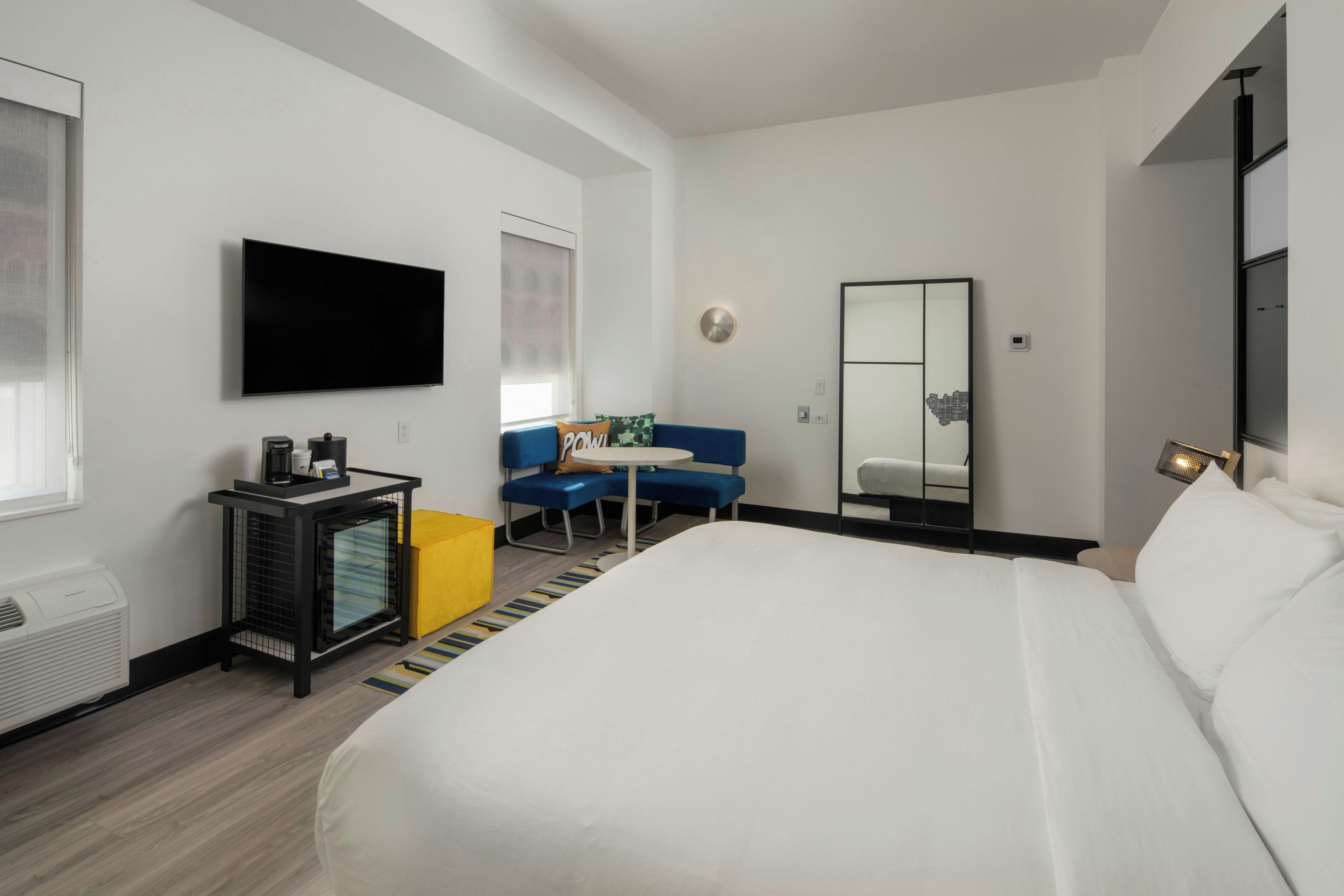 A hotel room with a large bed, a flat-screen TV mounted on the wall, a desk with a coffee machine, and a seating area with a blue sofa.