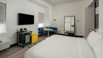 A hotel room with a large bed, a flat-screen TV mounted on the wall, a desk with a coffee machine, and a seating area with a blue sofa.
