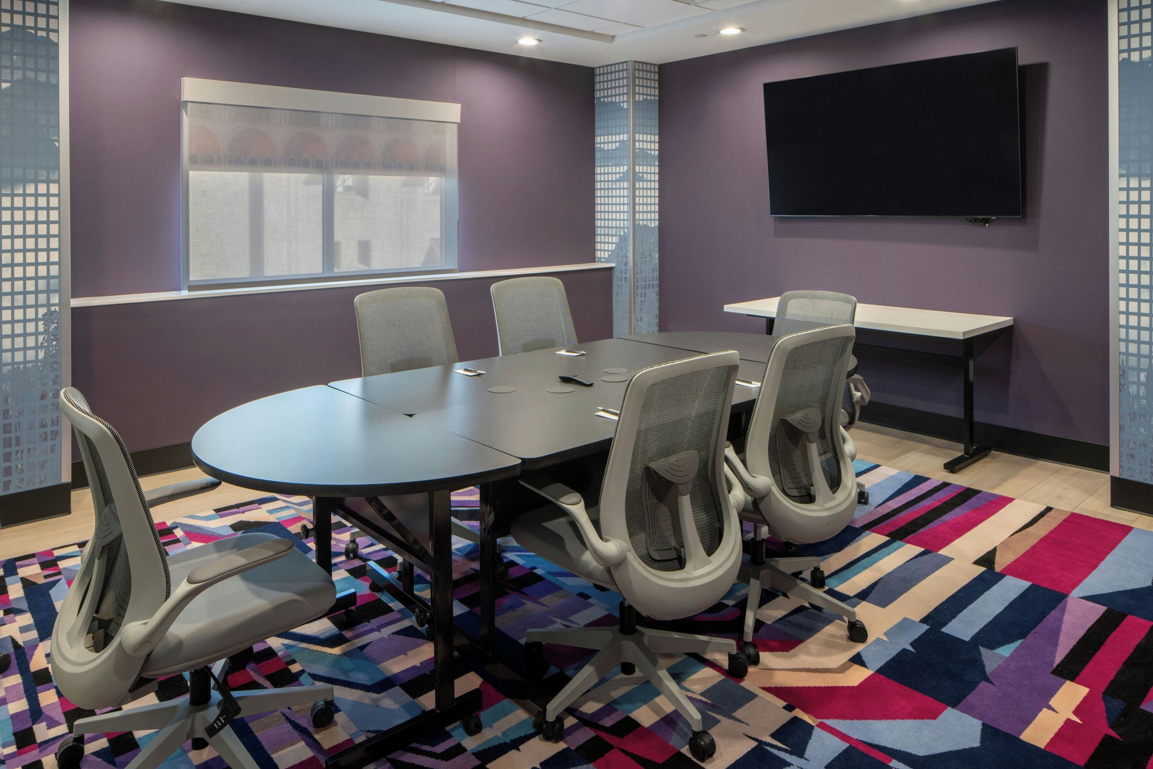 A conference room with a large table, six chairs, a projector screen, and a colorful rug.