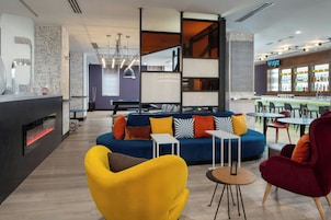 Un área de estar moderna con un sofá azul, un sillón amarillo y una silla roja. Hay una barra con un letrero de neón, y una mesa de billar al fondo.
