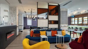 A modern lounge area with a blue sofa, yellow armchair, and red armchair. There is a bar with a sign reading "WXYZ" and a fireplace with a visible fire. - Aloft by Marriott Memphis Downtown (Memphis)