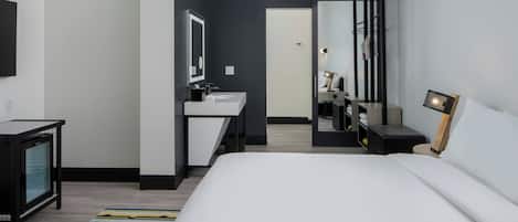 Habitación de hotel moderna con una cama grande, una mesita de noche, un espejo, un escritorio pequeño con una silla y un baño visible a través de una puerta abierta.