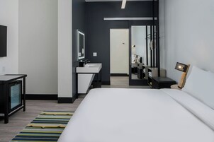Habitación de hotel moderna con una cama grande, una mesita de noche, un espejo, un escritorio pequeño con una silla y un baño visible a través de una puerta abierta.