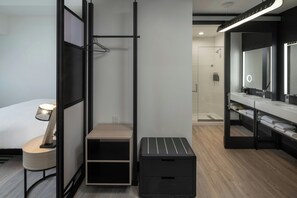 Ett modernt hotellrum med en säng, ett sängbord med en lampa, en svart förvaringsenhet, en träbänk, en svart pall och ett badrum med en stor spegel och ett handfat.
