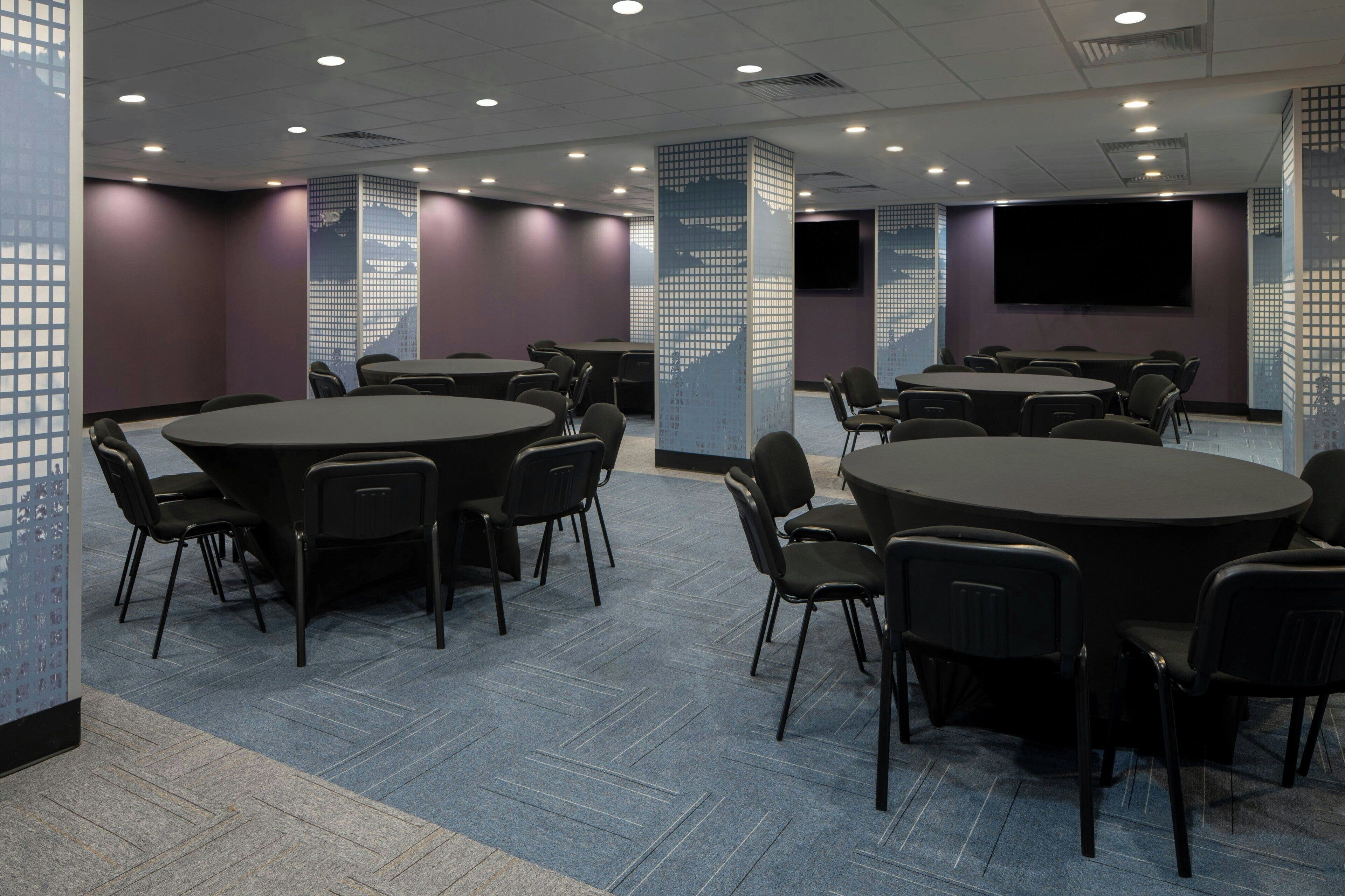 A conference room with multiple tables and chairs, a large screen, and a patterned wall.
