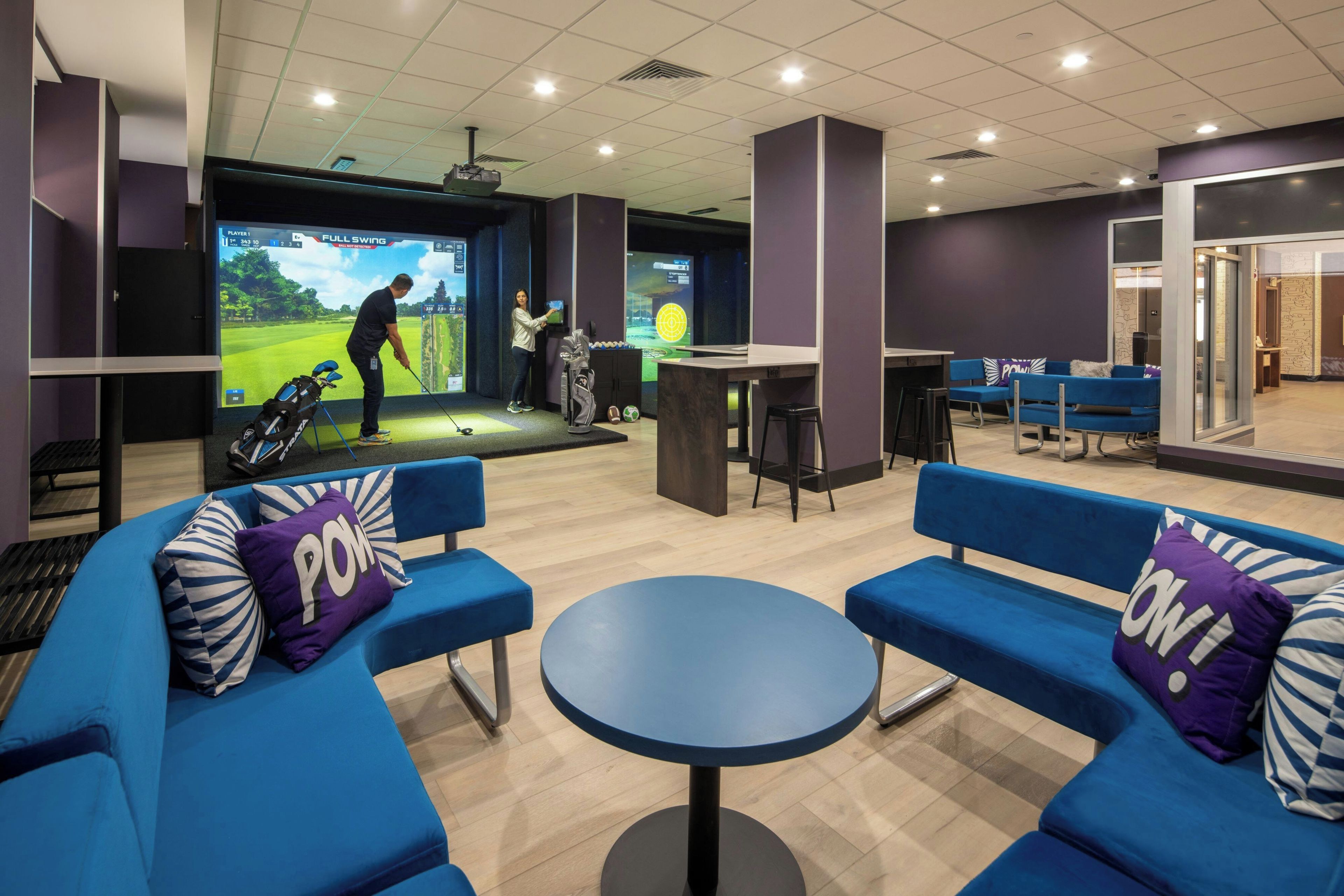 Un espacio interior moderno con asientos azules, una pantalla de proyección mostrando un partido de golf y una zona de bar.