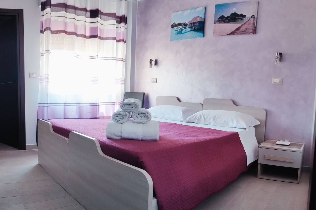 Un lit bien fait, avec une literie blanche et rose, une table de chevet sur laquelle se trouve une boîte de mouchoirs, et deux tableaux représentant des paysages côtiers, accrochés au mur.