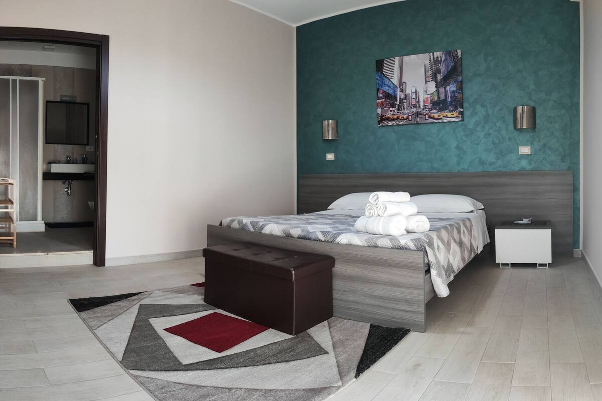 Une chambre d’hôtel moderne comprenant un lit, une petite table de chevet, une commode, un banc, une salle de bain avec un miroir et un lavabo, et un tableau représentant un paysage urbain accroché au mur.