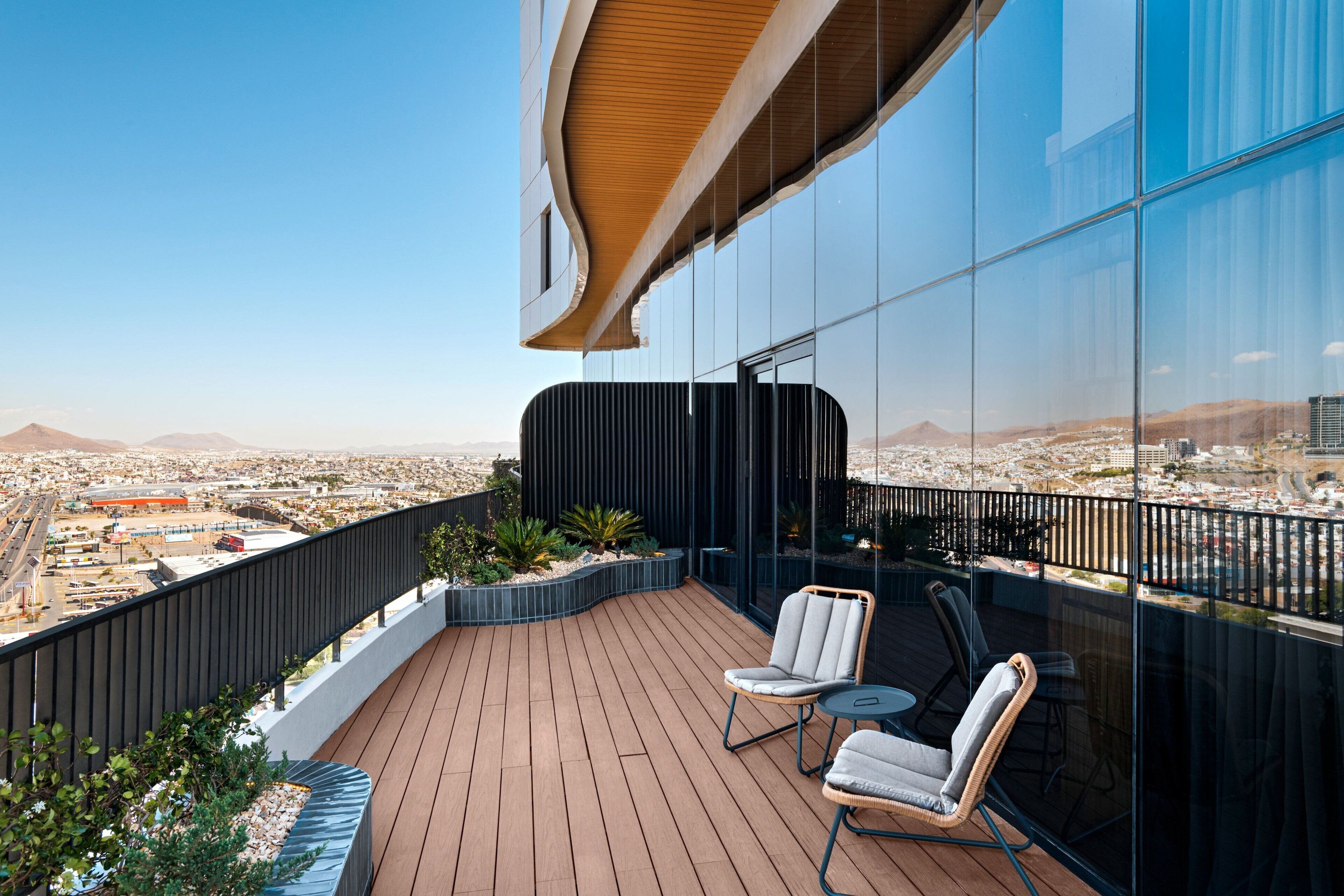 Un balcón moderno con suelo de madera, dos sillas con cojines y vistas a una ciudad y montañas al fondo.