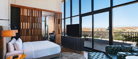 Quarto de hotel moderno com uma cama grande, televisão, varanda com vista para o deserto e área de estar com mobiliário de couro.