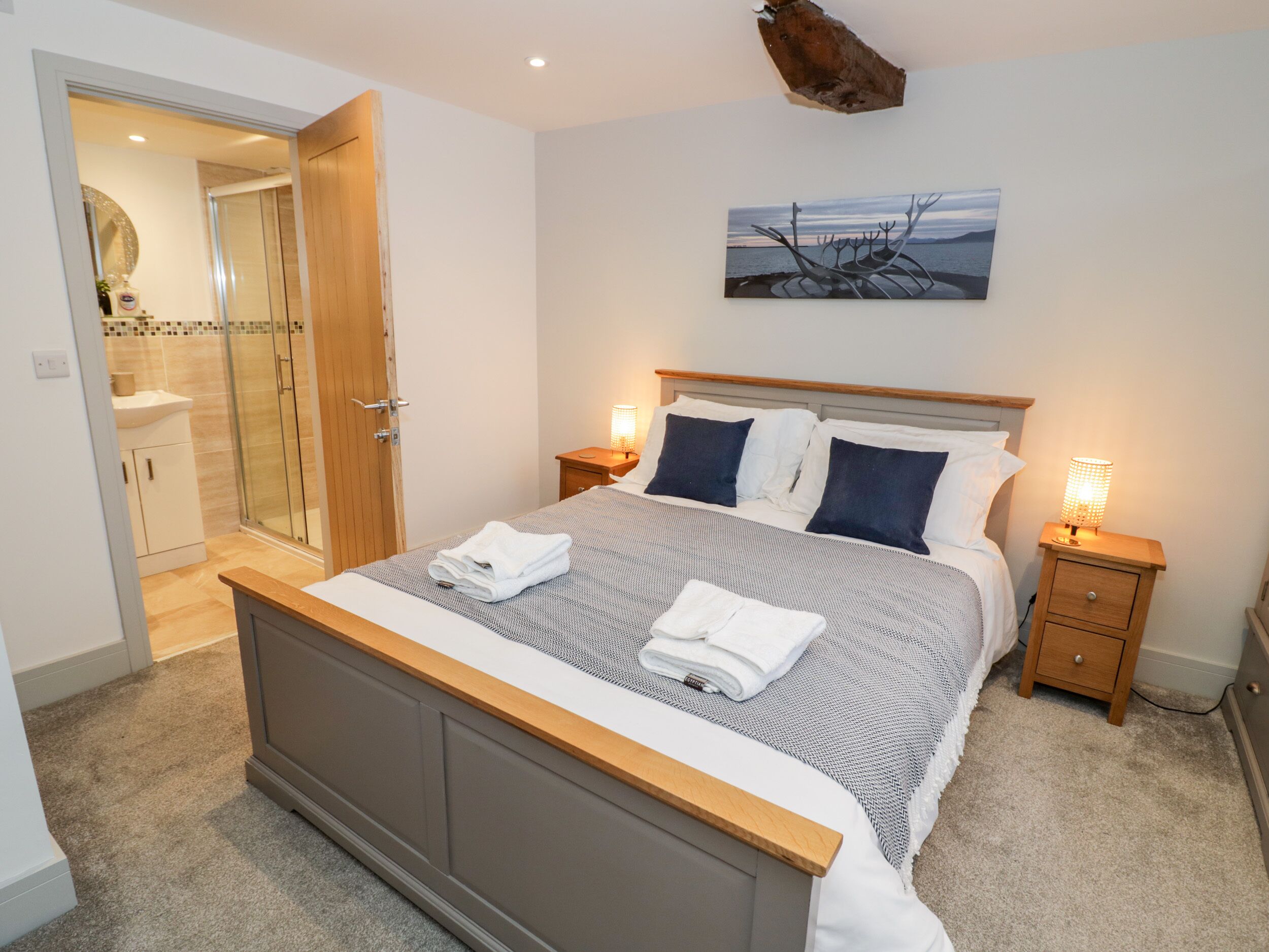 A bedroom with a bed, bedside tables, a nightstand, a painting, and a bathroom with a shower and toilet.