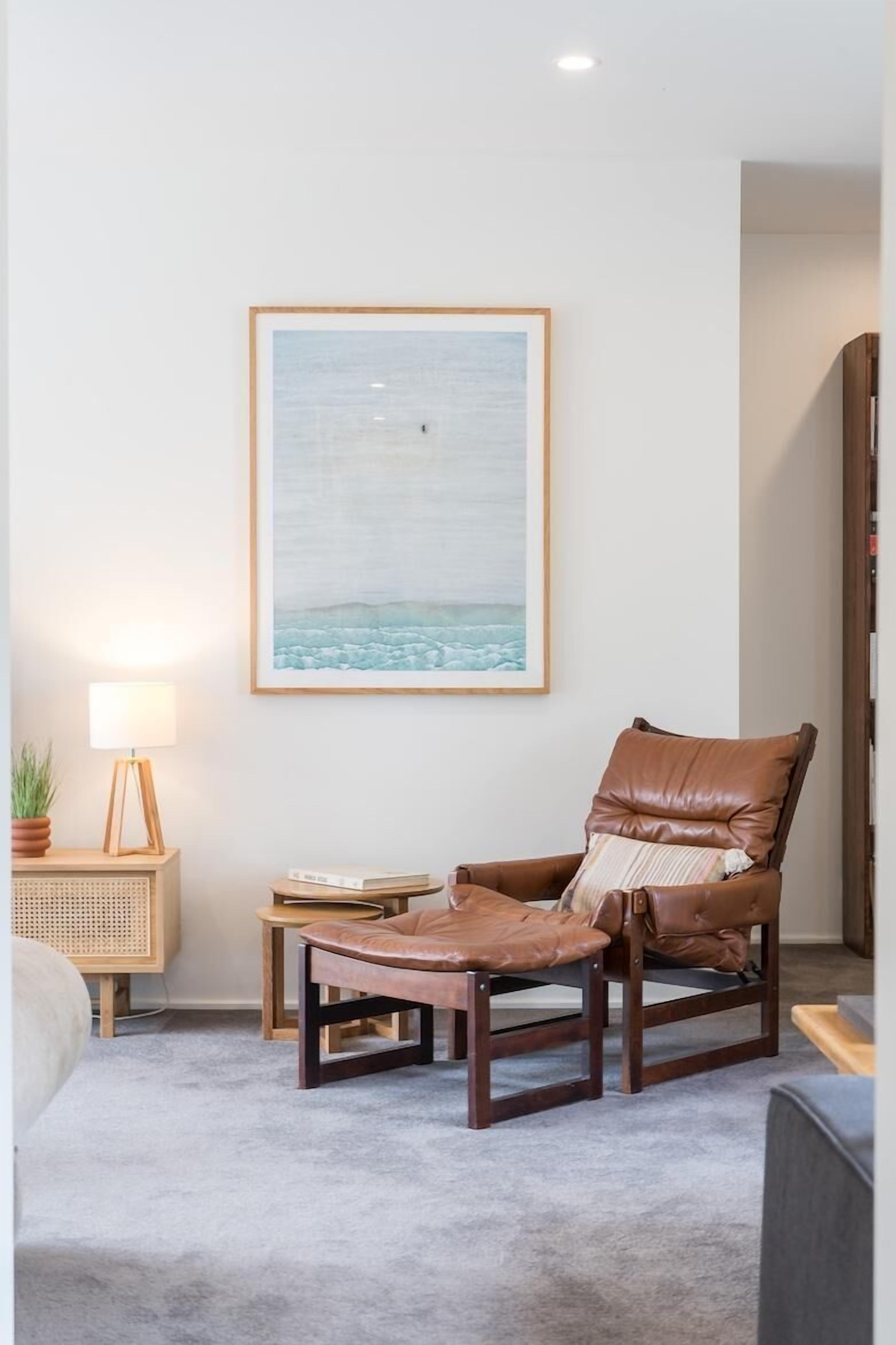 A living room with a brown leather armchair, a small wooden side table with a lamp, and a framed picture of a beach scene on the wall.