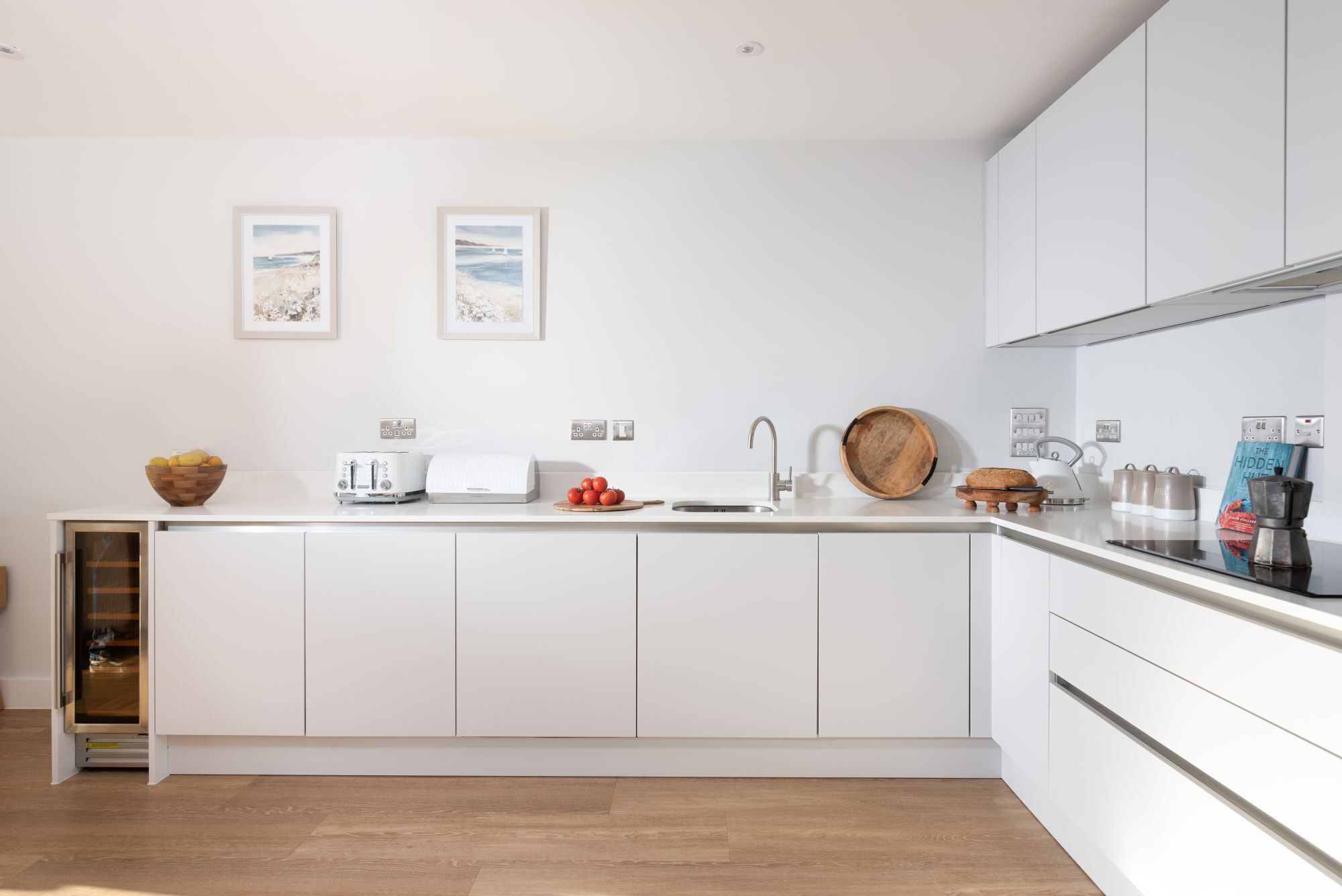 Een moderne keuken met witte kastjes, een ingebouwde oven en een groot schiereiland met een spoelbak. Twee ingelijste foto's hangen aan de muur, er staat een fruitschaal op het aanrecht en een waterkoker.