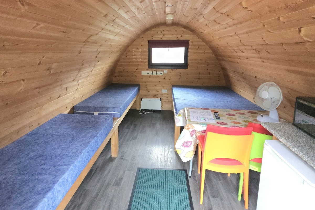 Une petite cabane en bois, chaleureuse et accueillante, avec deux lits simples, une petite table et un ventilateur.