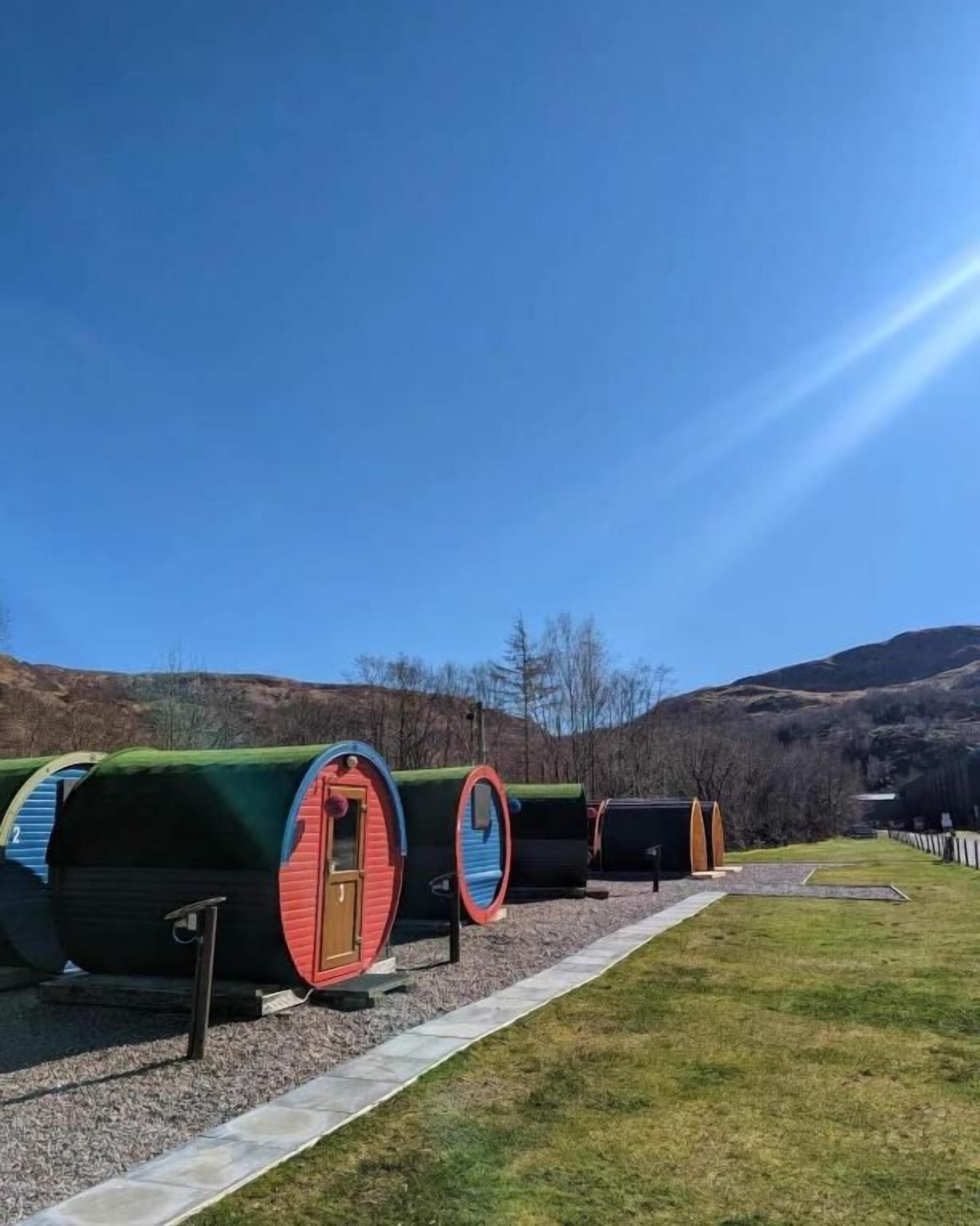 A row of cylindrical cabins with doors, set on a gravel path, surrounded by grass and hills.