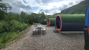 A series of cylindrical structures with red doors, possibly for sleeping or resting, set on a gravel path with picnic tables. - Glamping Pods (Kinlochleven)