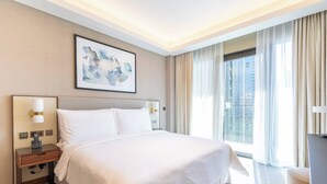 Une chambre d’hôtel avec un grand lit, une table de chevet avec une lampe, un bureau avec une chaise, une fenêtre avec des rideaux et un tableau encadré au mur.
