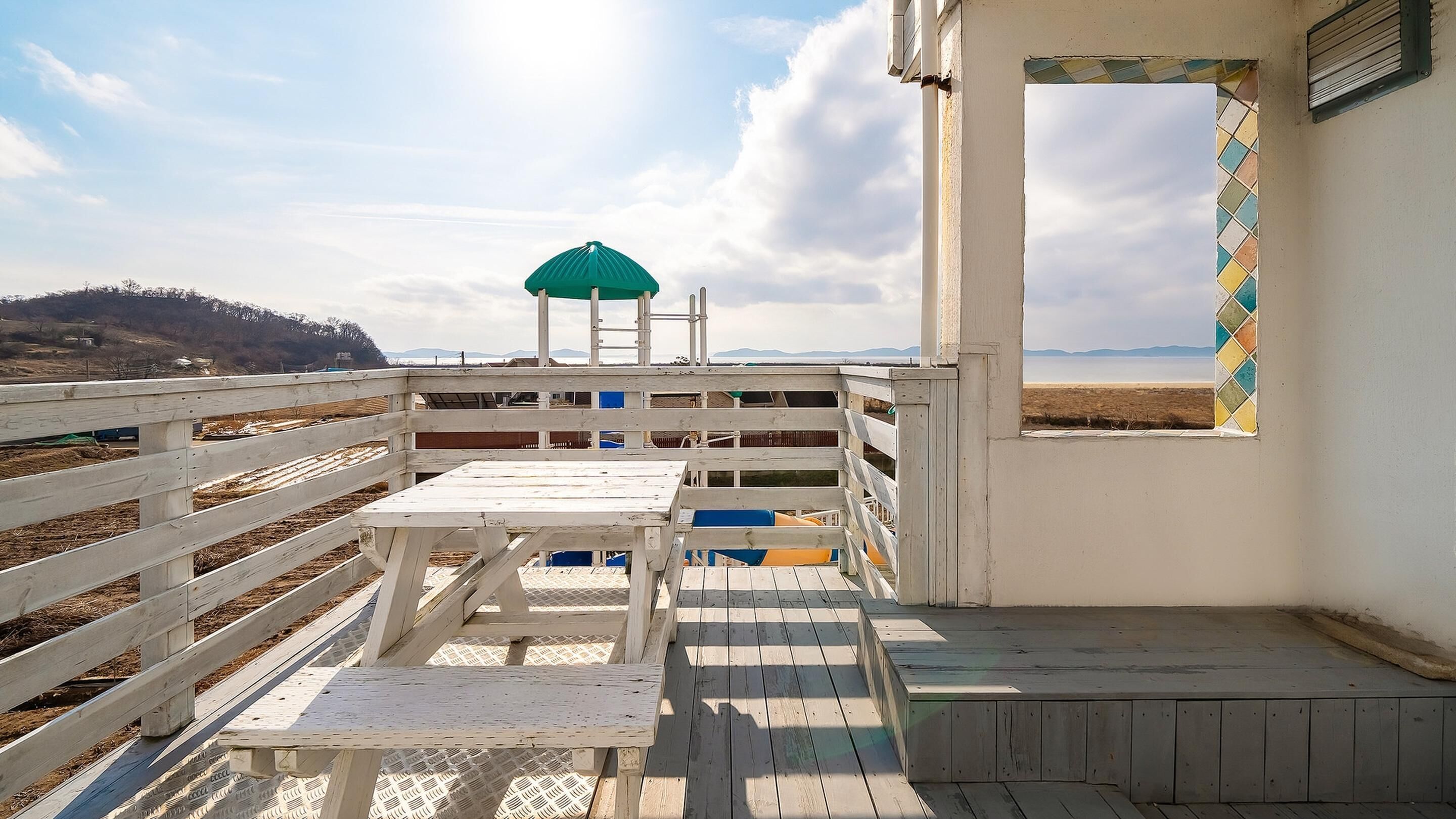 Une terrasse en bois avec une table et des chaises, un parasol vert et une vue sur la mer et les montagnes.