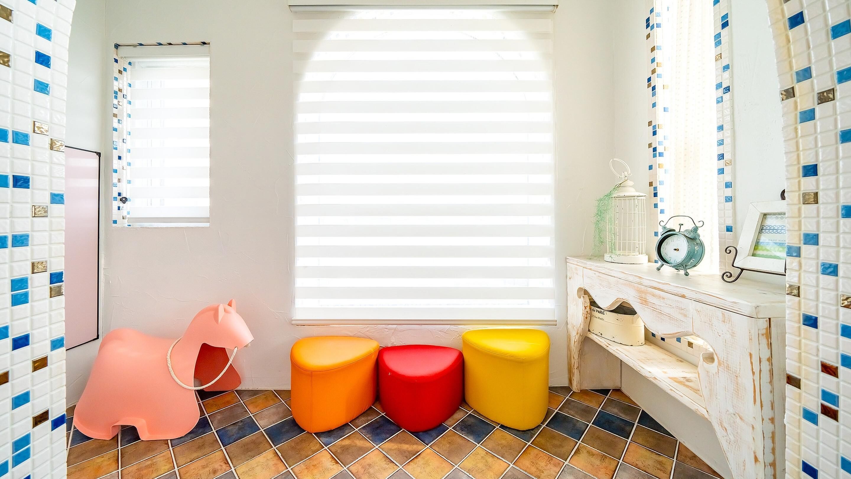 A bathroom with a white sink, a mirror, and a window with blinds. There are colorful stools and a pink chair.