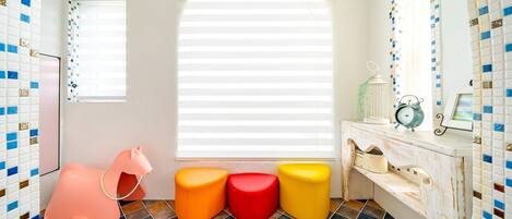 A bathroom with a white sink, a mirror, and a window with blinds. There are colorful stools and a pink chair.