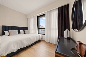 A hotel room with a bed, a desk, a chair, a window with curtains, and a view of buildings. - Luxurious 3-Bdr In King David Street with Parking (Jerusalem)